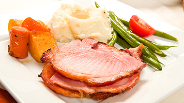Ham with sweet potatoes, mashed potatoes, and green beans and a tomato.